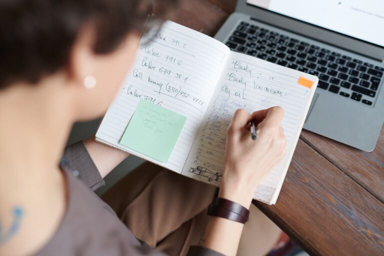 Person writing notes in a notebook next to an open laptop